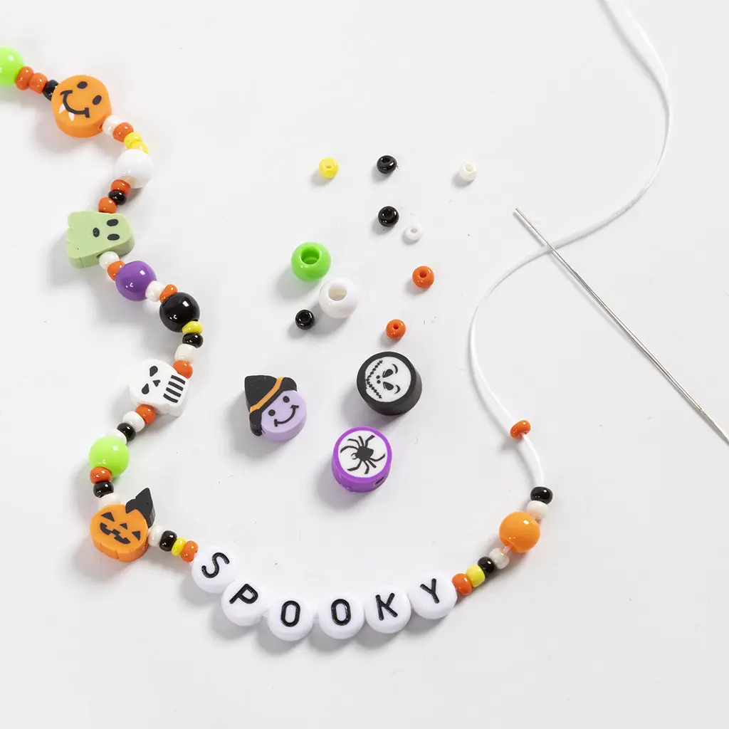 Een rijgdraad met een rij kleurrijke Halloween-kraaltjes en letterkraaltjes die het woord 'SPOOKY' vormen, met losse kraaltjes en een naald erbij op een witte ondergrond.