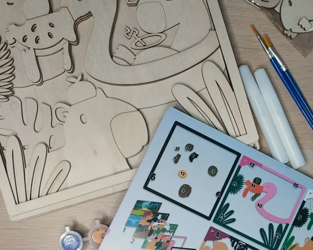 Bovenaanzicht van een houten lasergestanst capybara-kunstpakket met meerdere lagen, een instructiekaart met genummerde verfpotjes en een flamingo-afbeelding, twee penselen en witte lijmtubes