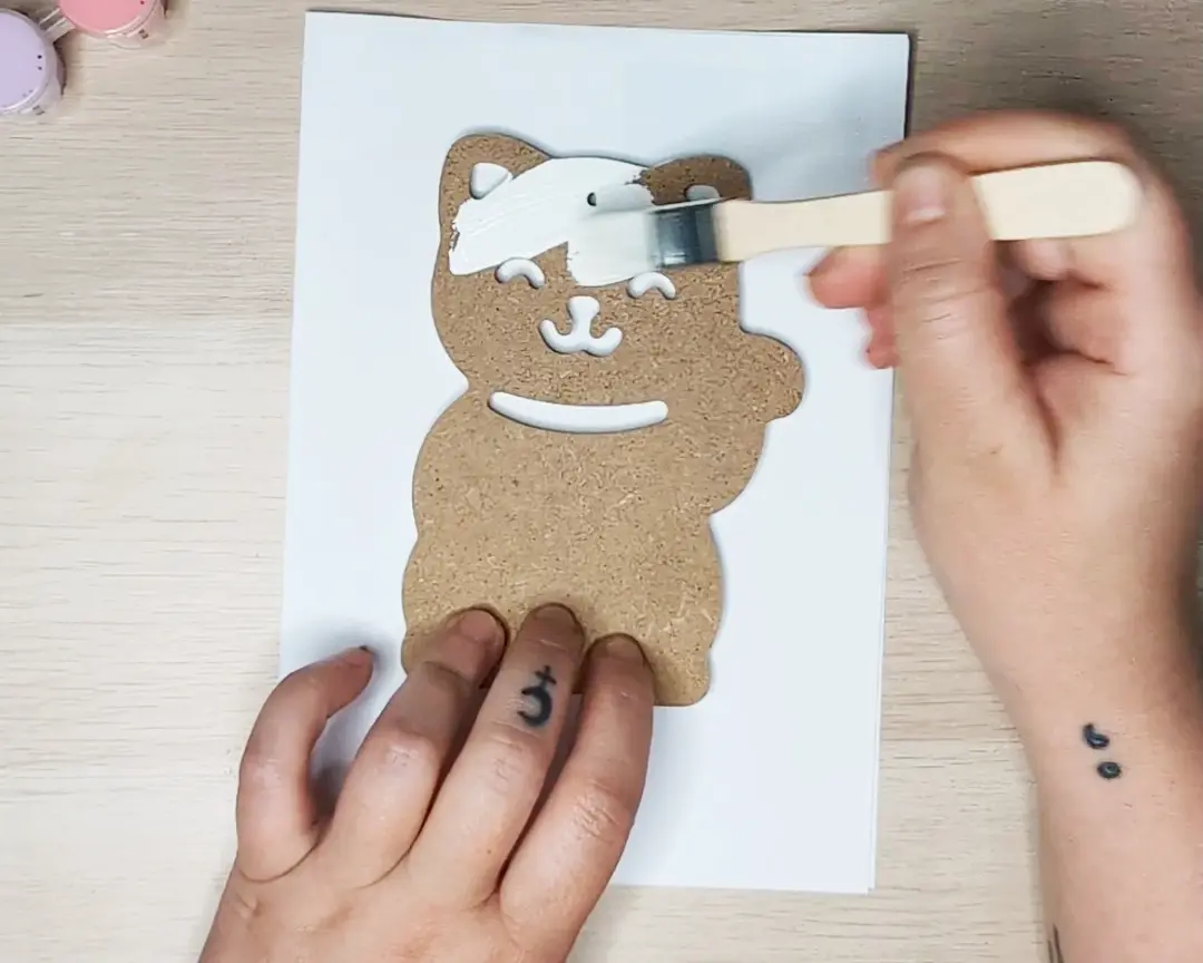 Handen die een mdf maneki-neko figuur vasthouden en wit beschilderen met een plat kwastje, op een witte ondergrond.