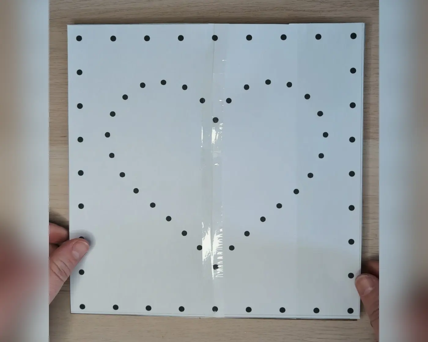 Twee handen houden een wit papier met zwarte stippen in een hartvorm, vastgehouden met tape in het midden