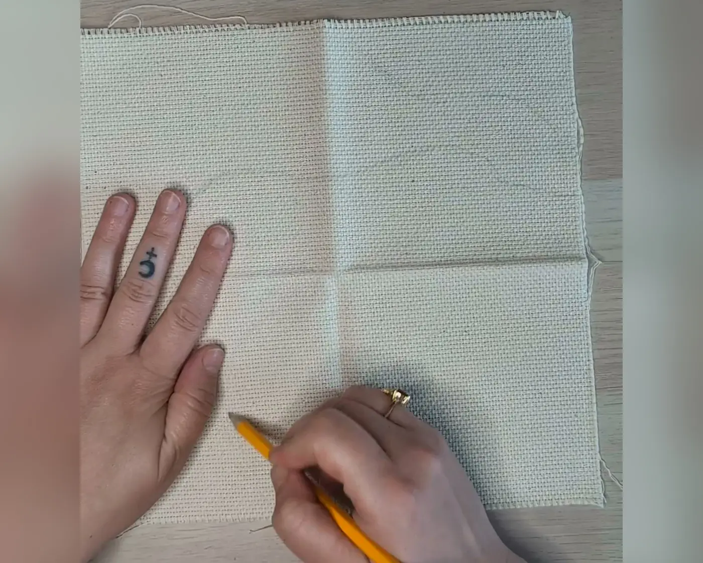 Hand die met een potlood een ontwerp overtekent op beige punch needle stof, uitgespreid op een houten ondergrond