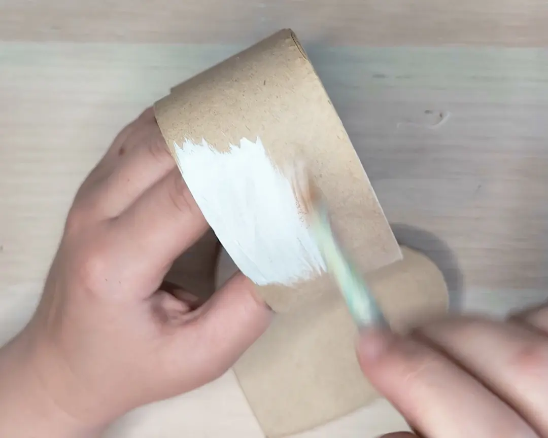 Twee handen schilderen de binnenkant van een wc-rolletje wit met een kwastje.