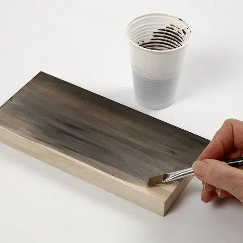 Een hand verft een rechthoekige houten plank met een transparante grijze verf of beits met behulp van een platte kwast. Op de achtergrond staat een witte plastic beker met verf.