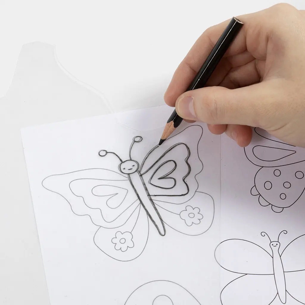 Een hand overtrekt met een zwart potlood de contouren van een vlindertekening op een vel transparante krimpfolie.