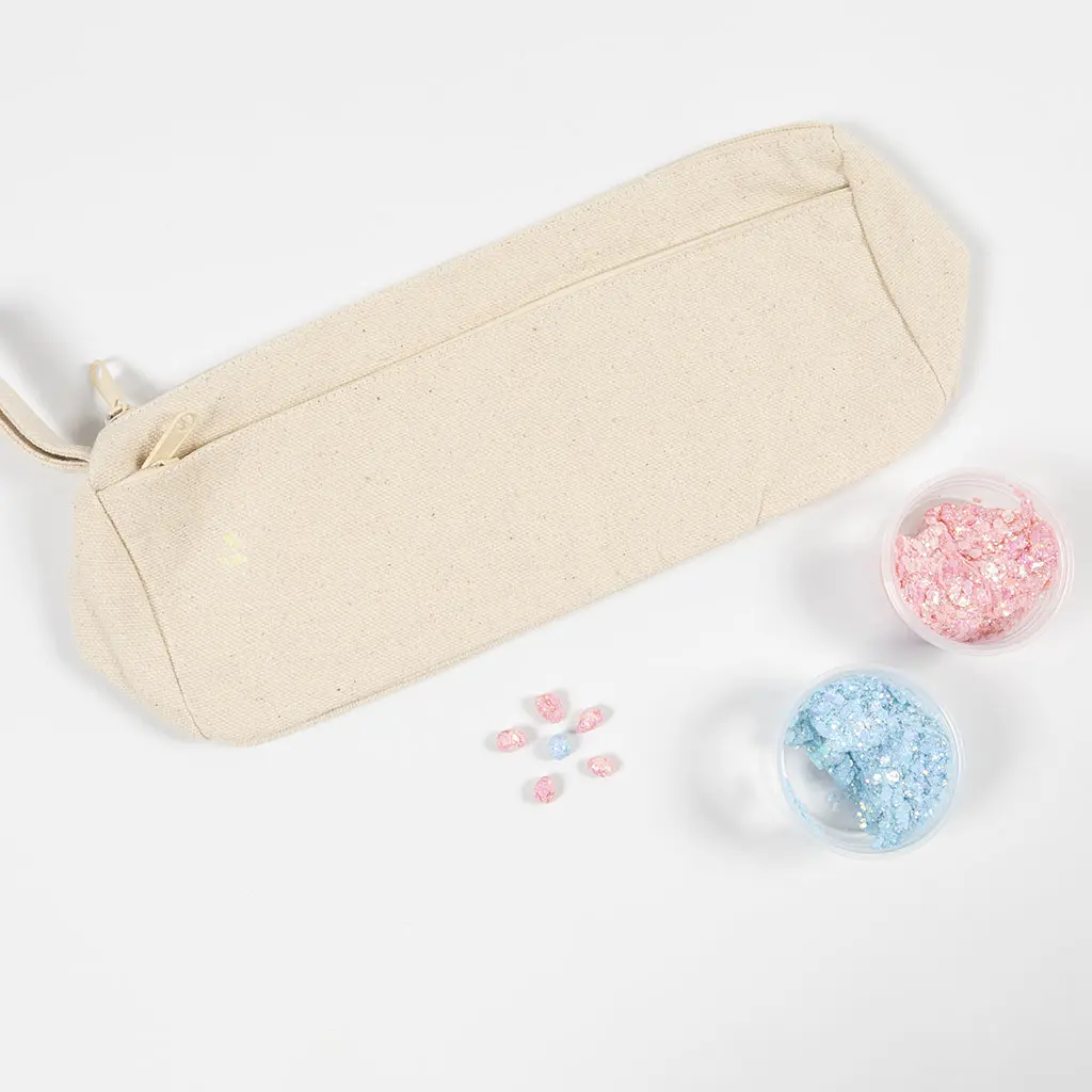 Een naturel canvas etui met rits, naast twee open potjes chunky glitter in roze en lichtblauw en enkele losse glitterstenen.