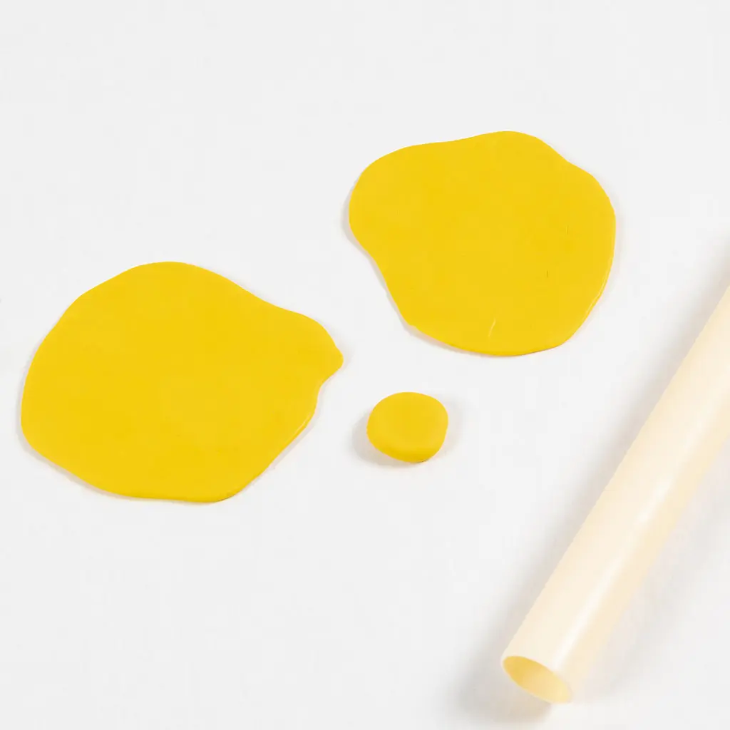 Bovenaanzicht van twee platgerolde, onregelmatige cirkels van gele polymeerklei op een witte achtergrond. Hiertussen ligt een klein, rond bolletje gele klei en rechtsonder is een beige plastic roller zichtbaar.
