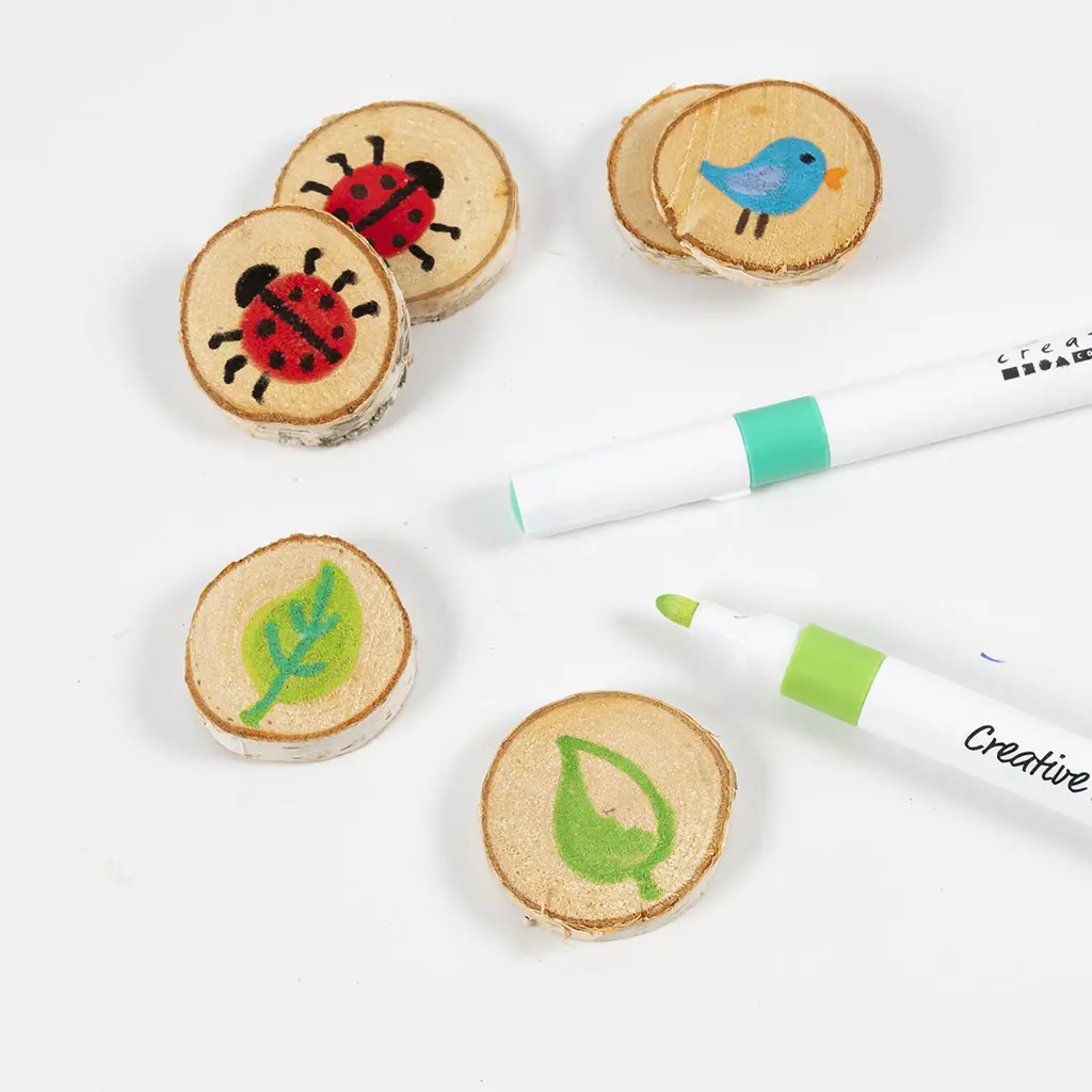 Bovenaanzicht van verschillende kleine, ronde boomschijfjes die zijn beschilderd met natuurmotieven, waaronder lieveheersbeestjes, een blauw vogeltje en groene bladeren. Er liggen twee Creative Multi Markers (één turquoise en één lichtgroen) bij op de witte tafel.