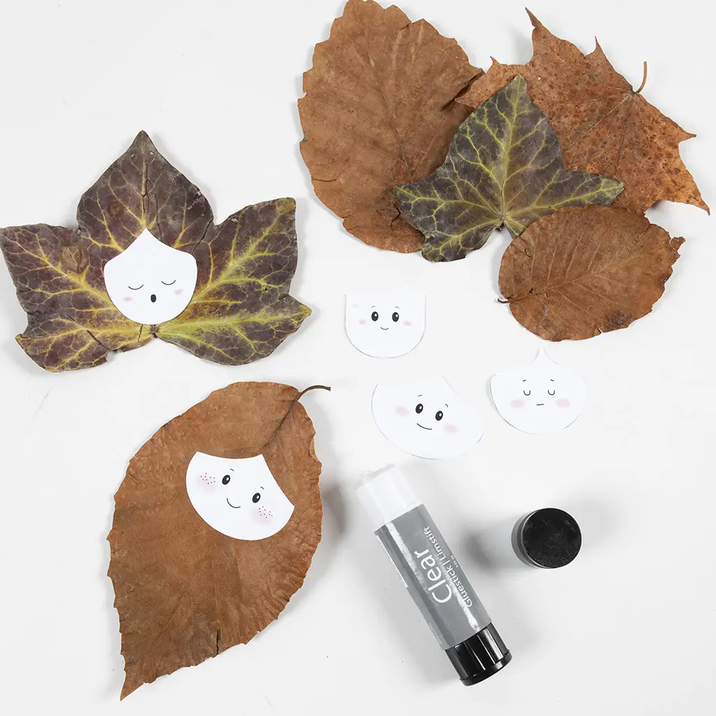 Gedroogde herfstbladeren, kleine getekende papieren gezichtjes en een pritt lijmstift uitgestald op een witte achtergrond.