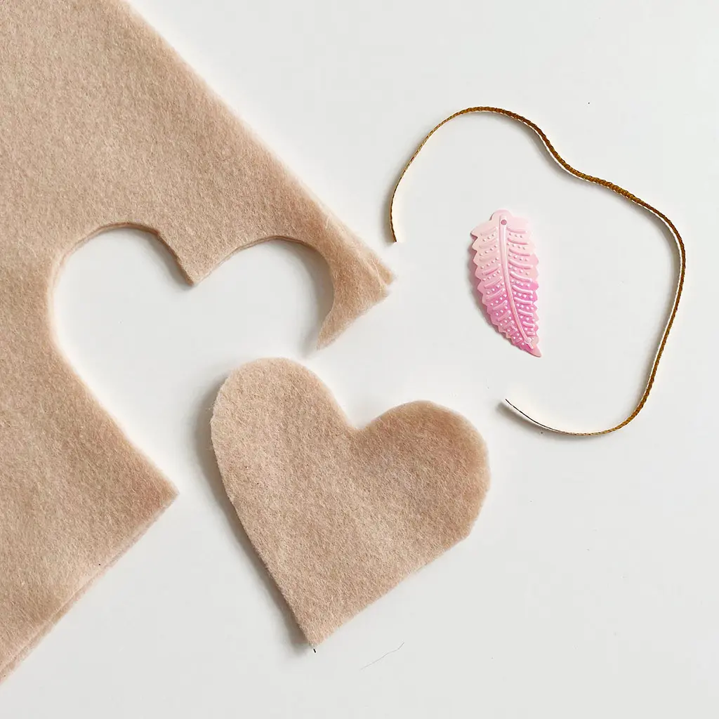 Uitgeknipte roze vilten hartjes op een witte achtergrond, samen met goudkleurig lint en een roze glimmende paillet in de vorm van een veer.