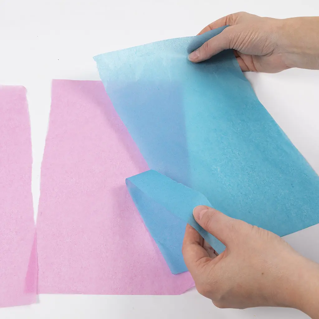 Handen scheuren verticale stroken uit vellen gekleurd vloeipapier in roze en blauw.