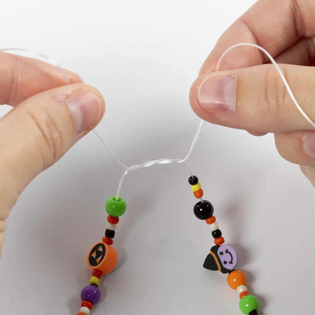 Twee handen die de uiteinden van de witte elastische draad kruisen om een eerste knoop in het armbandje te leggen.