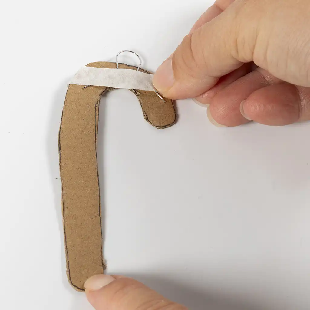 Bovenaan de uitgeknipte kartonnen zuurstok wordt een klein metalen ophangoogje vastgezet met een strookje schilderstape.