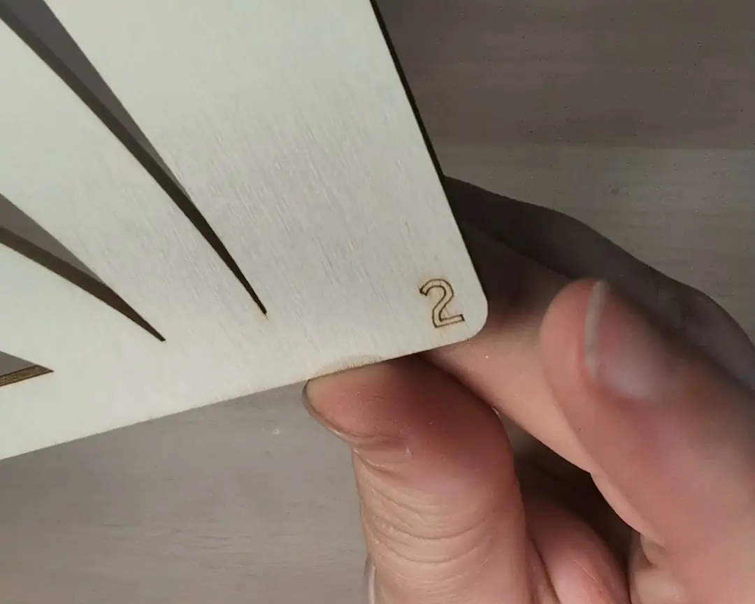 Detailopname van een hand die een houten laagplaat vasthoudt met het cijfer 2 ingegraveerd in de hoek.
