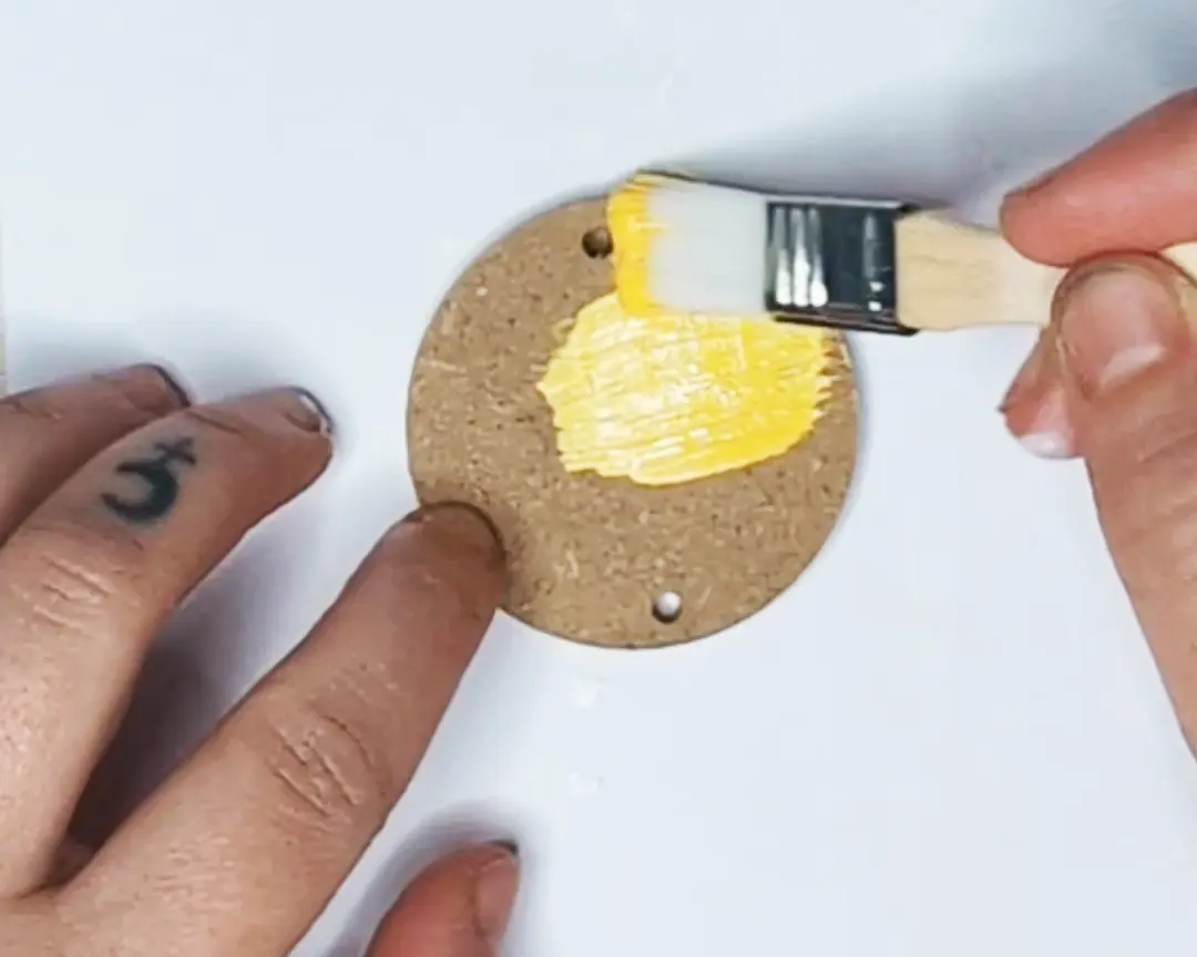 Hand die een rond MDF schijfje met twee gaatjes beschildert met gele verf en een plat kwastje.