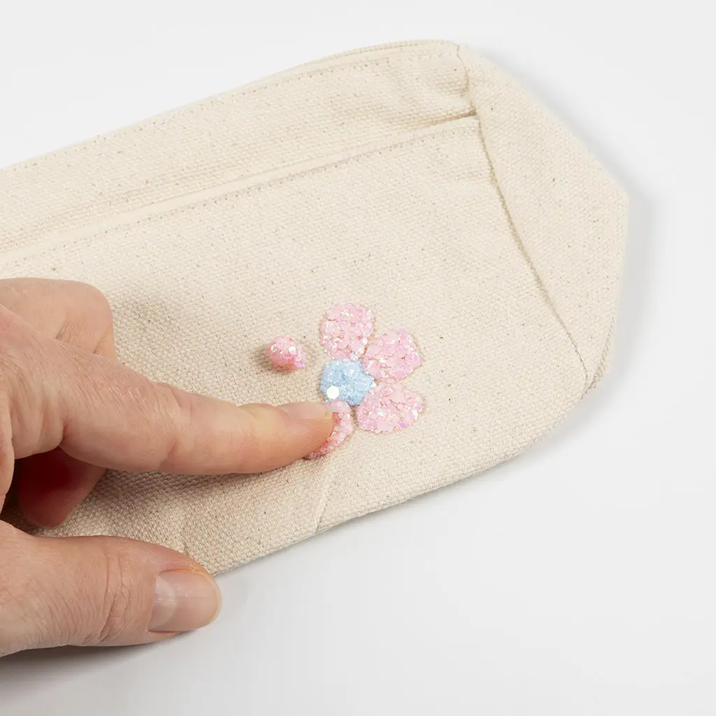 Een hand drukt een lichtblauw glitter hartje in het midden van een roze glitter bloemetje op een naturel canvas etui.
