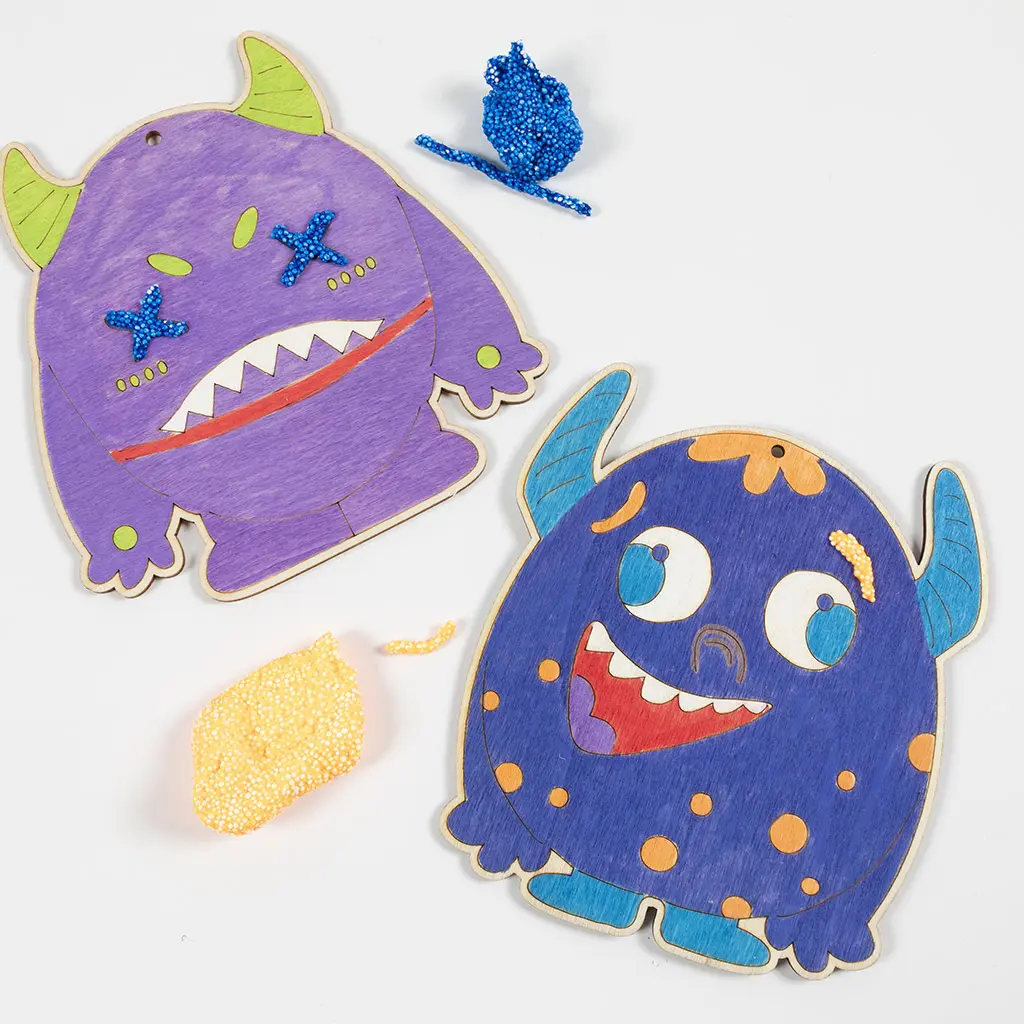 Twee ingekleurde houten monsters waarbij details worden versierd met Foam Clay. Het paarse monster heeft blauwe klei op zijn kruisogen en het blauwe monster heeft gele klei op zijn wenkbrauwen en oranje stippen op zijn lijf.