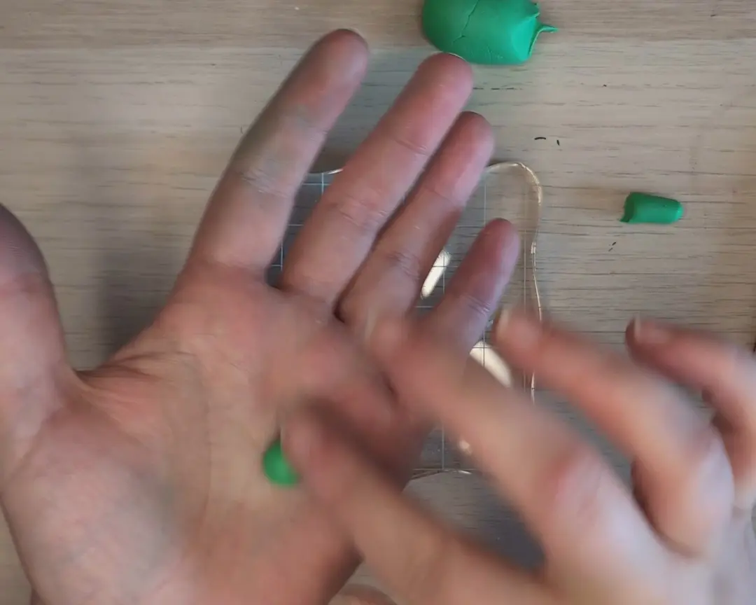 Een geopende handpalm waarin een klein, gladgerold groen bolletje klei ligt. Op de houten achtergrond liggen nog een groter stuk groene klei en een klein groen rolletje.