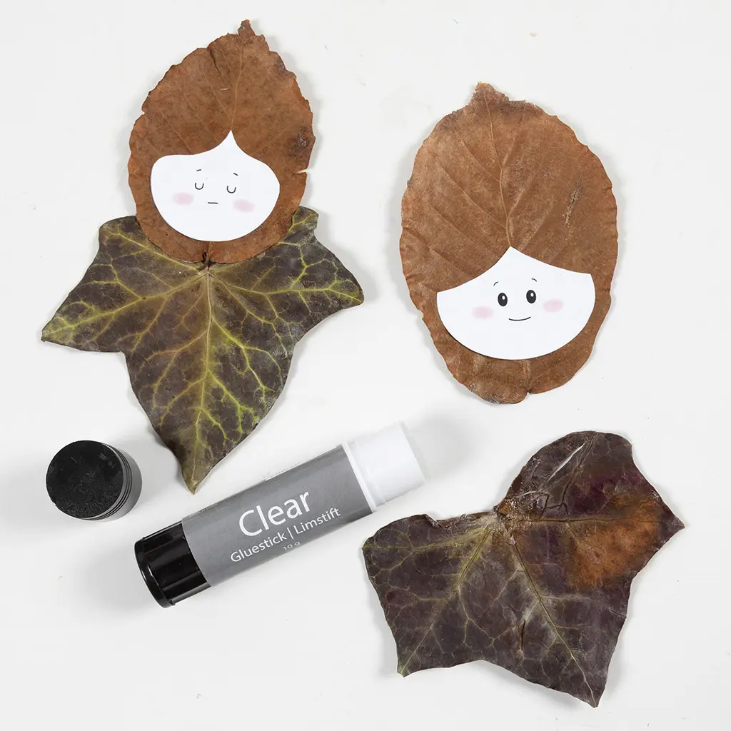 Twee gemaakte bladerfiguren met opgeplakte papieren gezichtjes en een open lijmstift op een witte ondergrond.