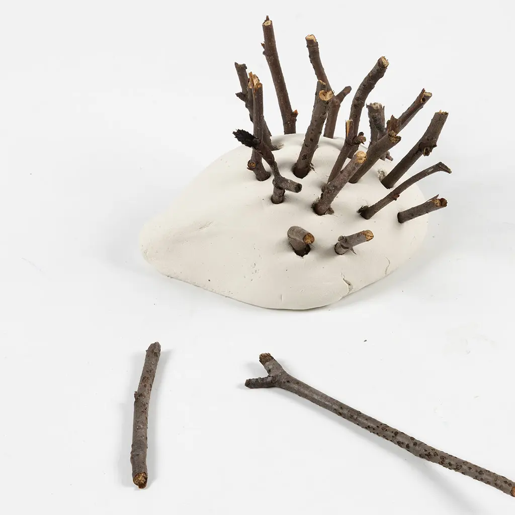 Een figuurtje van witte klei waarin korte boomtakjes en houtstokjes zijn gestoken als egelstekels.