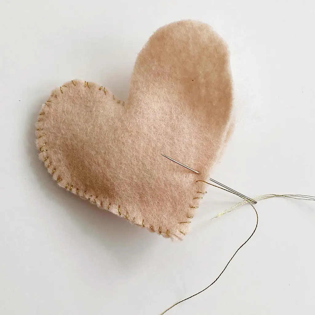 Detailopname van een roze vilten hartje dat langs de randen dichtgenaaid wordt met goudkleurig garen en een naald.