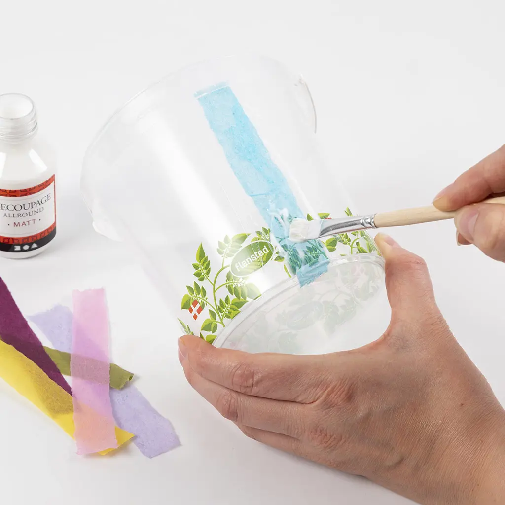 Een blauwe strook vloeipapier wordt met een platte kwast en decoupagelijm verticaal op een transparant plastic emmertje geplakt.