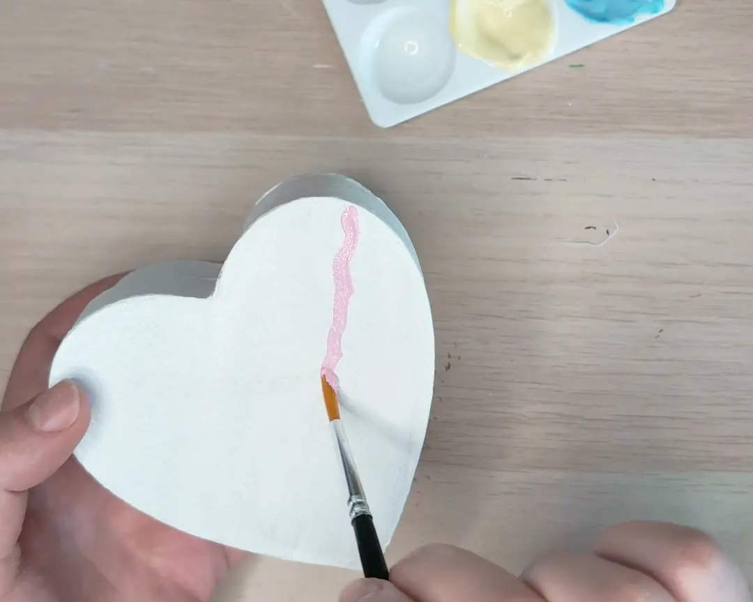 Een hand schildert golvende roze lijnen op een wit geschilderd hartvormig deksel met een fijn penseel, met een verfpalet op de achtergrond.
