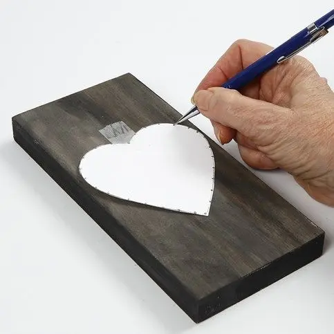 Een hand gebruikt een blauwe pen om stippen te zetten langs de rand van een papieren hart dat met tape op de donker geverfde houten plank is geplakt.