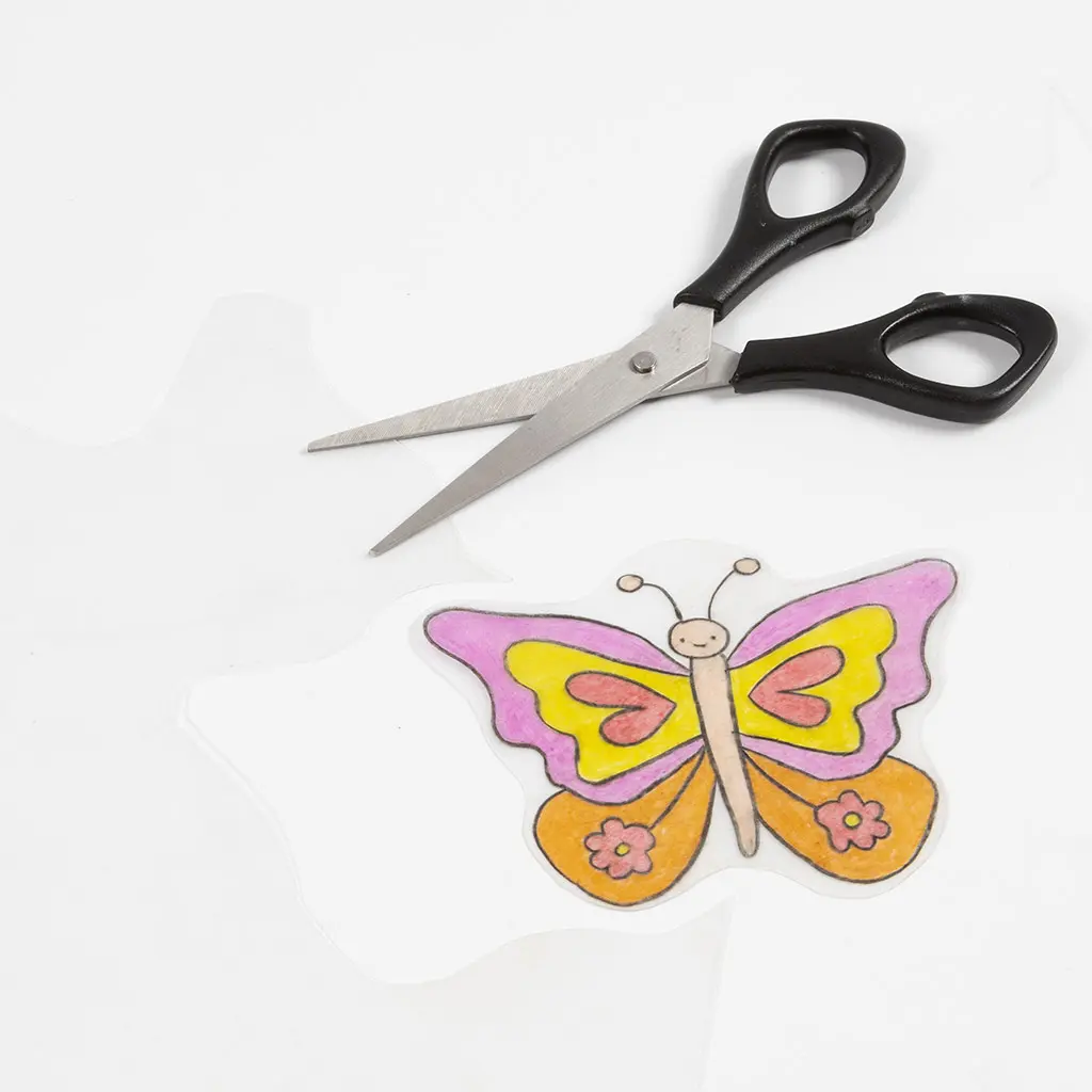 De ingekleurde vlinder is grof uitgeknipt uit de folie. Daarboven ligt een metalen hobbyschaar met zwarte handvaten.
