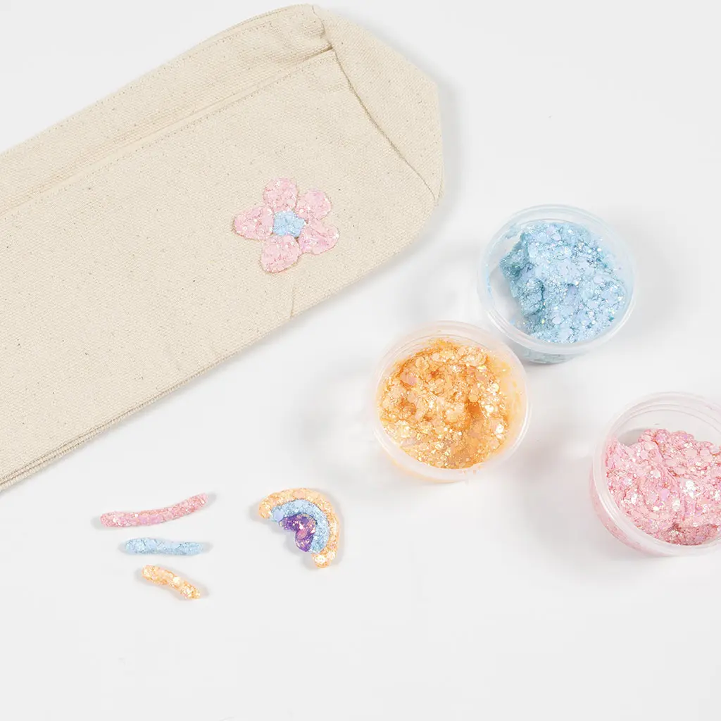Een naturel canvas etui met een roze glitter bloemetje, naast drie open potjes chunky glitter in lichtblauw, goud en roze, met losse glittersymbolen waaronder een regenboogje.