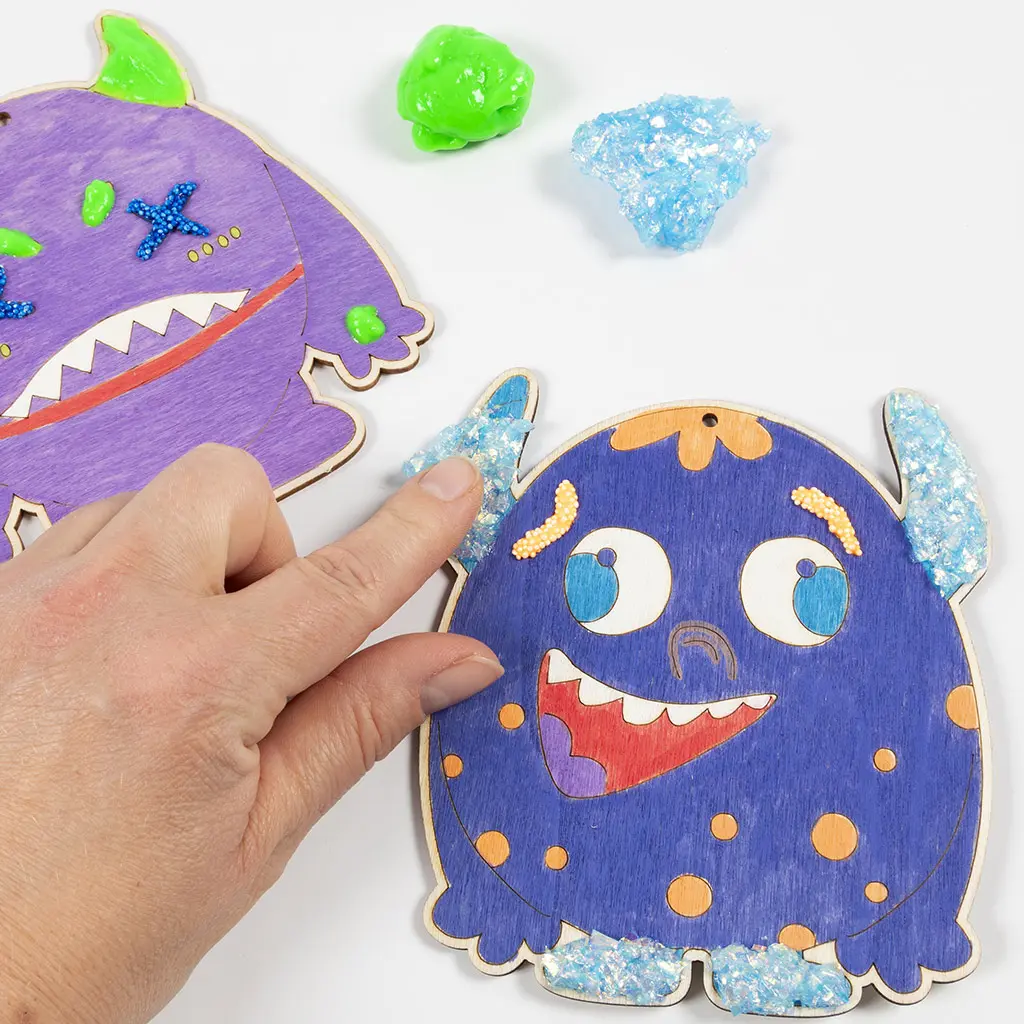 Een hand brengt glinsterende, blauwe glittergel aan op de hoorns van het blauwe houten monster. Op de achtergrond heeft het paarse monster felgroene klei op zijn hoorns gekregen.