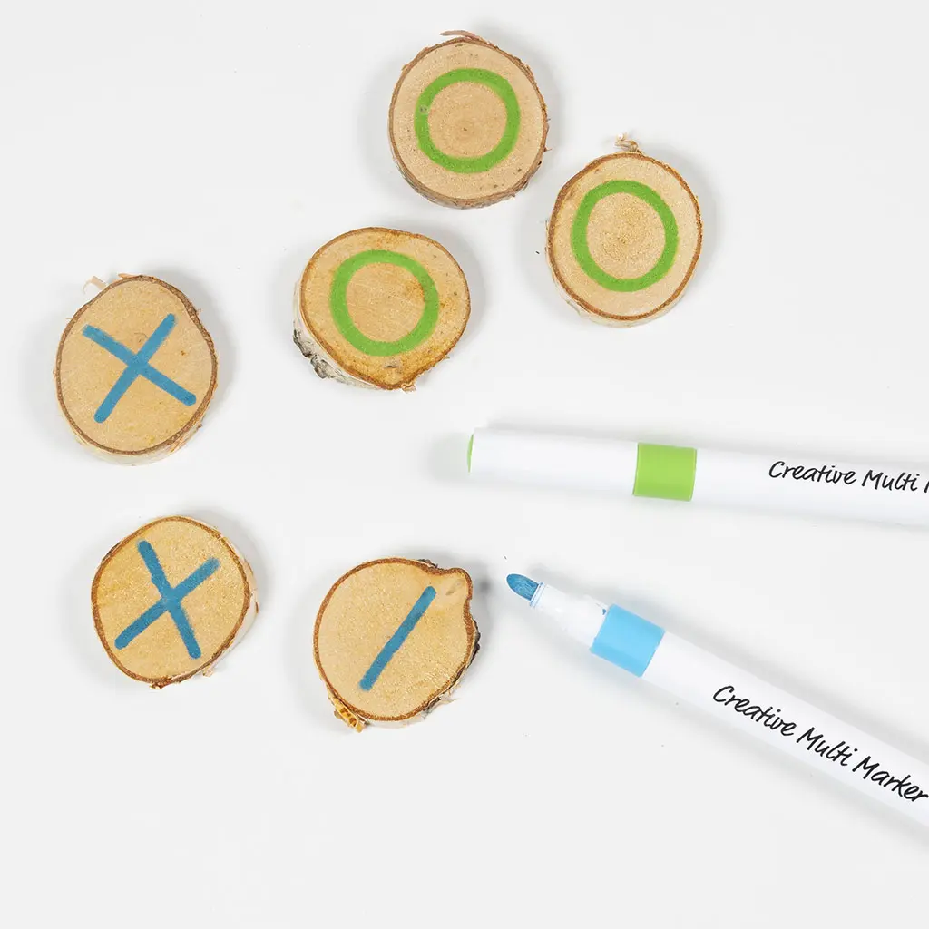 Een spelset in wording waarbij boomschijfjes worden gemarkeerd voor een spelletje boter-kaas-en-eieren. Drie schijfjes hebben een groen cirkel-symbool (O) en drie schijfjes hebben een blauw kruis-symbool (X). Er liggen een groene en een blauwe Creative Multi Marker naast.