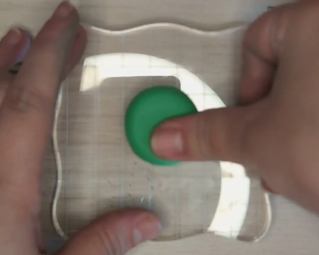 Een duim drukt een groen bolletje polymeerklei plat op een transparant acrylaatblok met ruitjespatroon op een houten ondergrond.