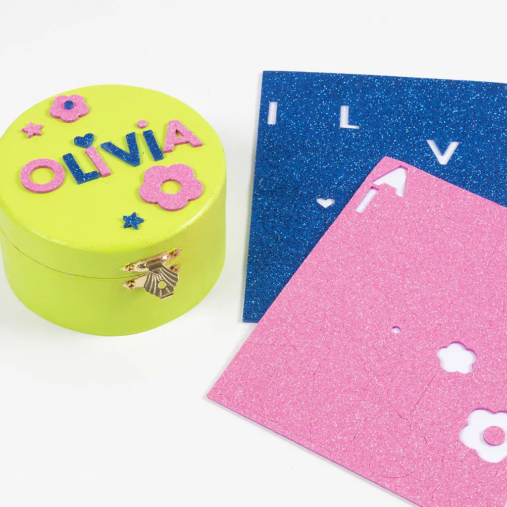 Een geel rond kistje met de naam Olivia erop, samengesteld uit foam letters in roze en blauw glitter, versierd met bloemen en sterren. Ernaast liggen vellen glitterfoam in blauw en roze waaruit letters en vormen zijn uitgesneden.