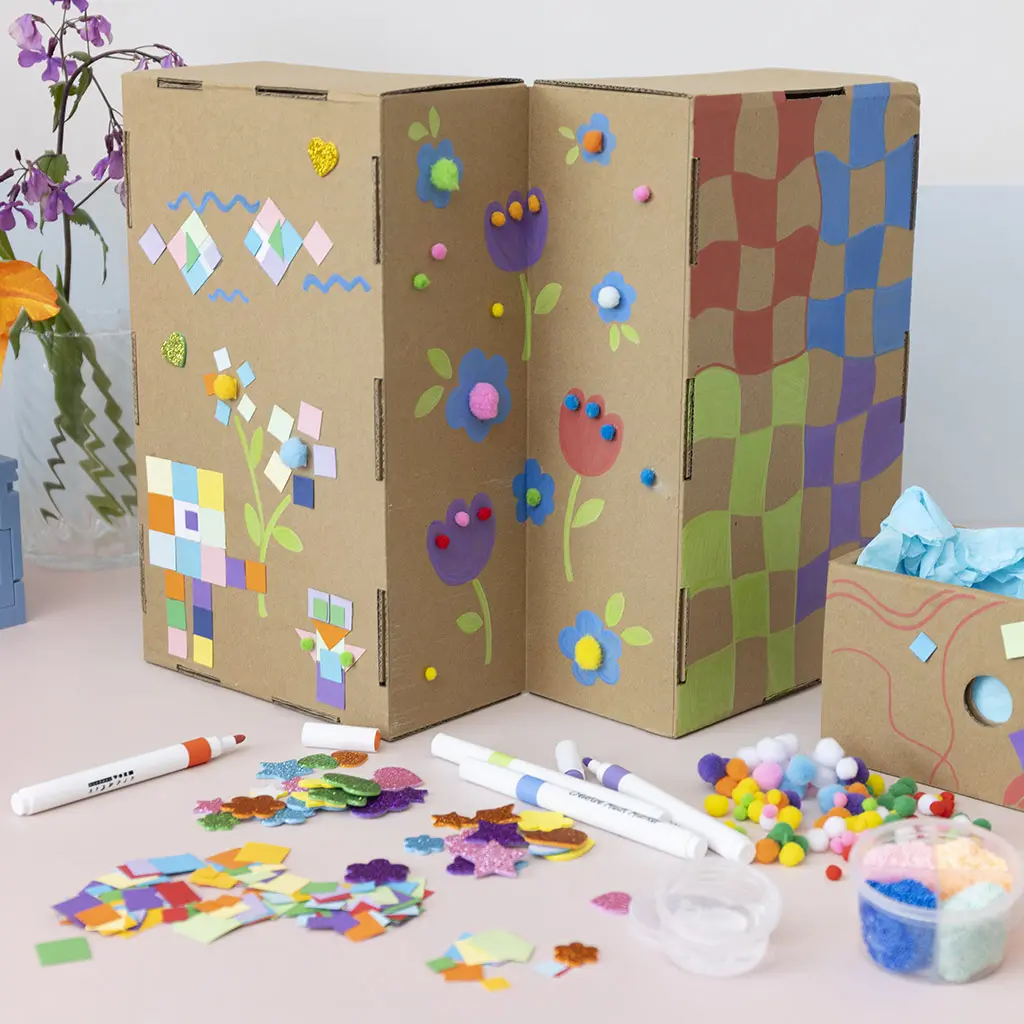 Achterzijde van een opklapbare kartonnen knutselkast versierd met geschilderde bloemen, blokpatronen en gekleurde stickers op een tafel met knutselbenodigdheden.