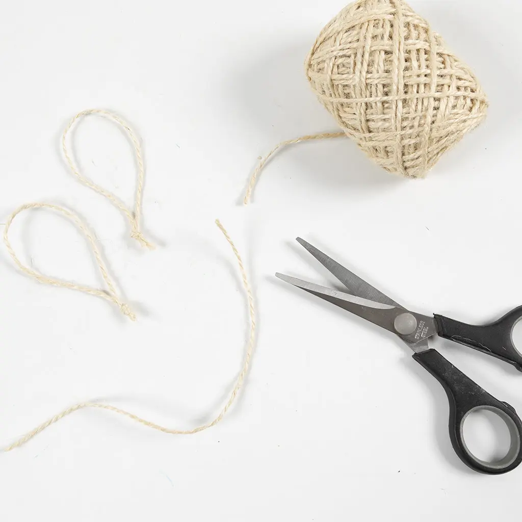 Een bol touw, een schaar en twee geknoopte lusjes van jute touw op een witte tafel.