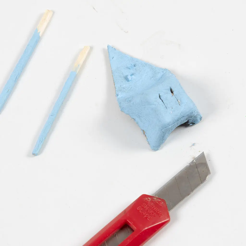Een precisiemesje naast een blauw geverfd kartonnen eierdoosdeel met ingesneden sleufjes en twee blauwe stokjes.