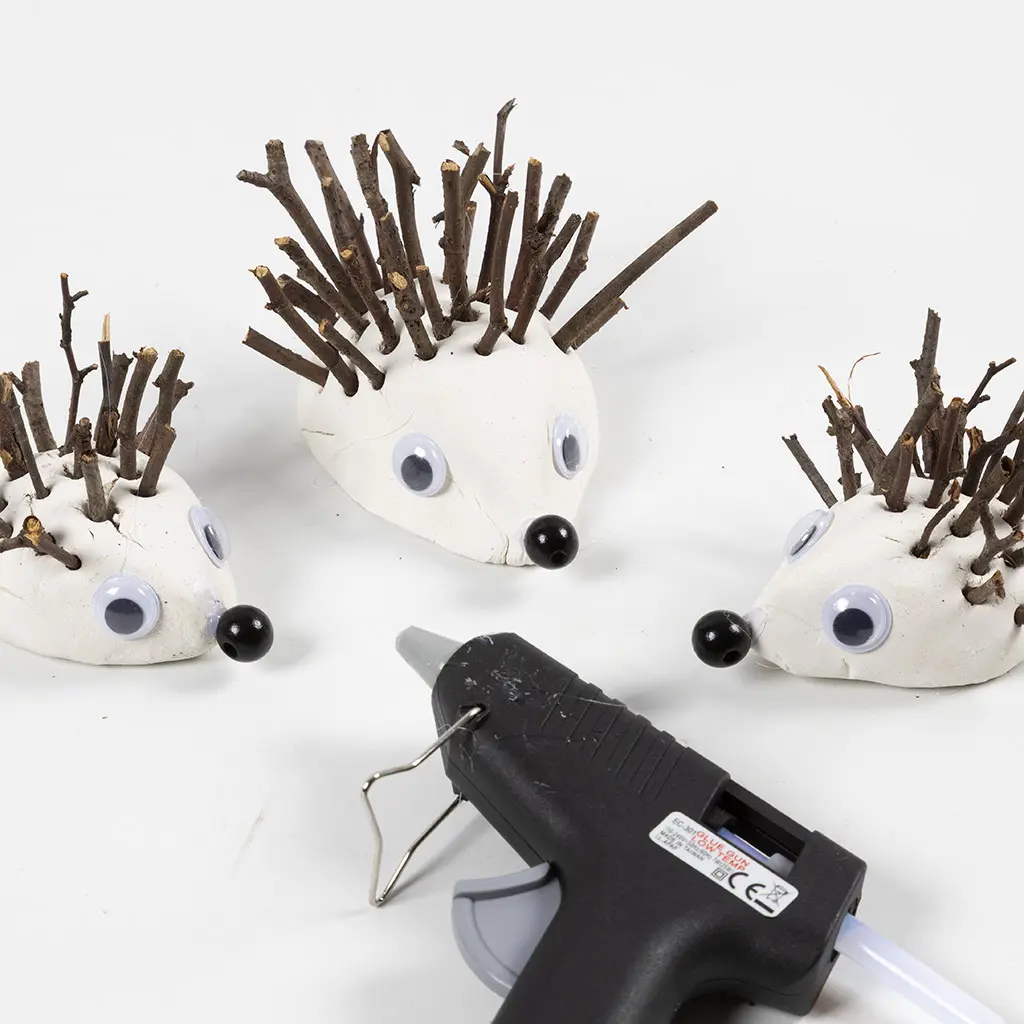 Drie handgemaakte kleie gels met takjes als stekels, giechelogen en een zwarte kraal als neus, met een lijmpistool op de voorgrond.