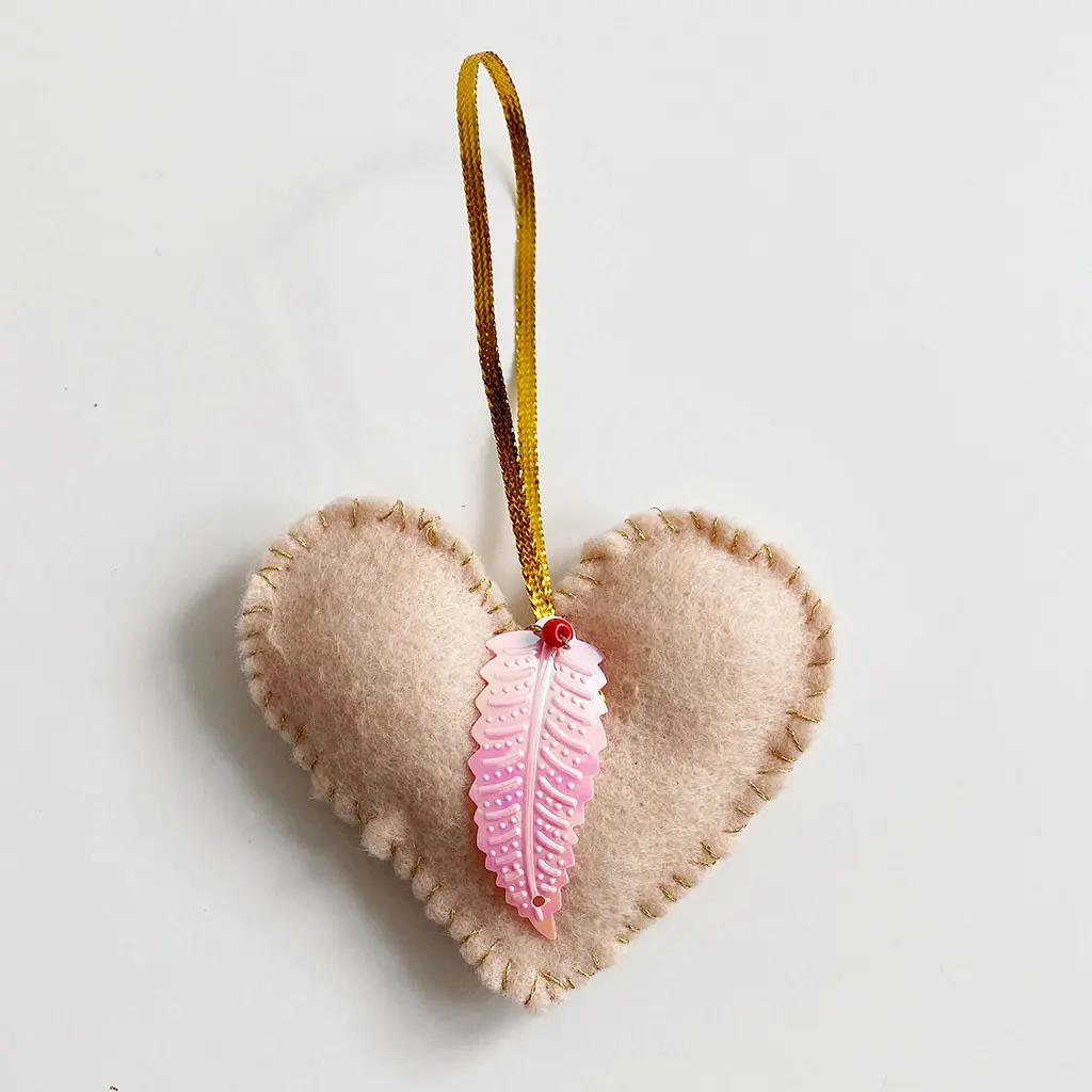 Een afgewerkt roze vilten hartjeshanger met gouden stiksels langs de rand, een gouden ophanglus en een roze veervormige paillet op het voorpand.