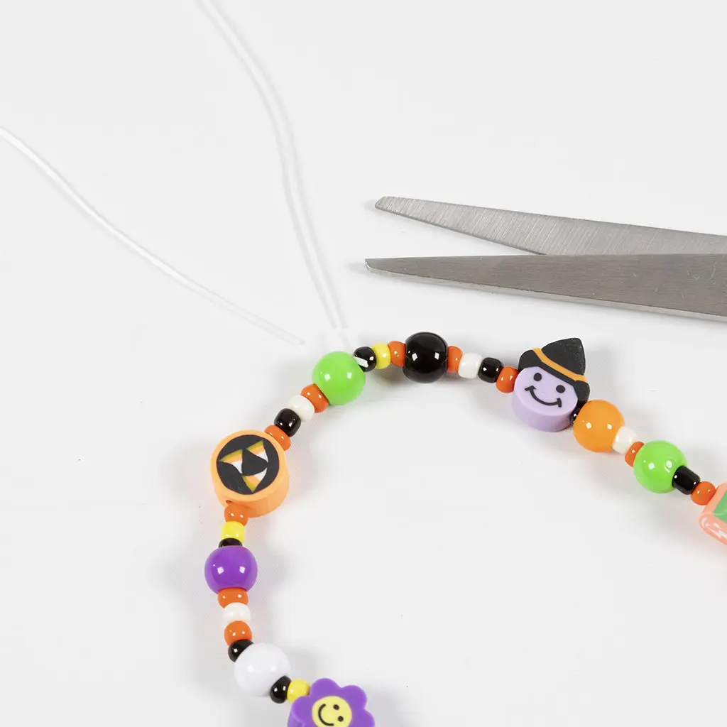 Een schaar die de overtollige uiteinden van de witte elastische draad vlak bij de knoop van het afgemaakte armbandje afknipt.