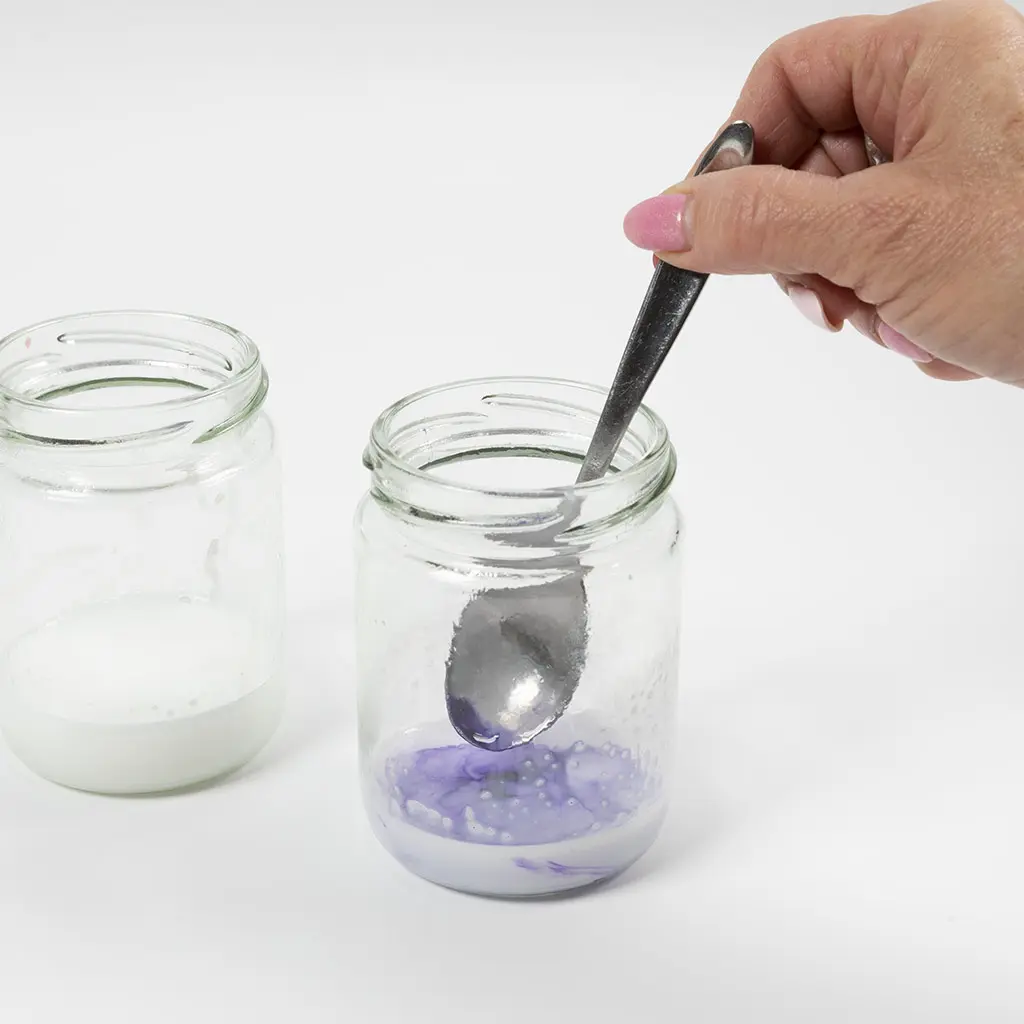 2 glazen spotten met gesmolten zeep, 1 wit en 1 lila. In de pot met lila zeep zit een lepel die het mengsel roert
