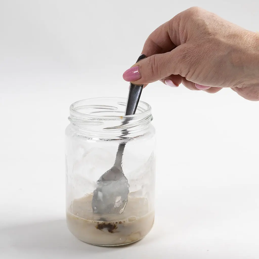 1 glazen pot met gesmolte bruine zeep. In de pot zeep zit een lepel die het mengsel roert