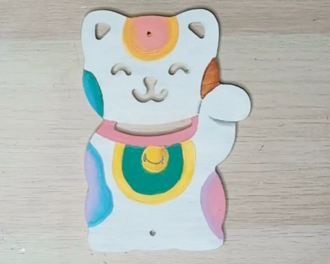 Volledig beschilderde MDF maneki-neko in pastelkleuren met roze halsband, gele en groene munt op de buik, en kleurrijke vlekken in blauw, paars en roze.