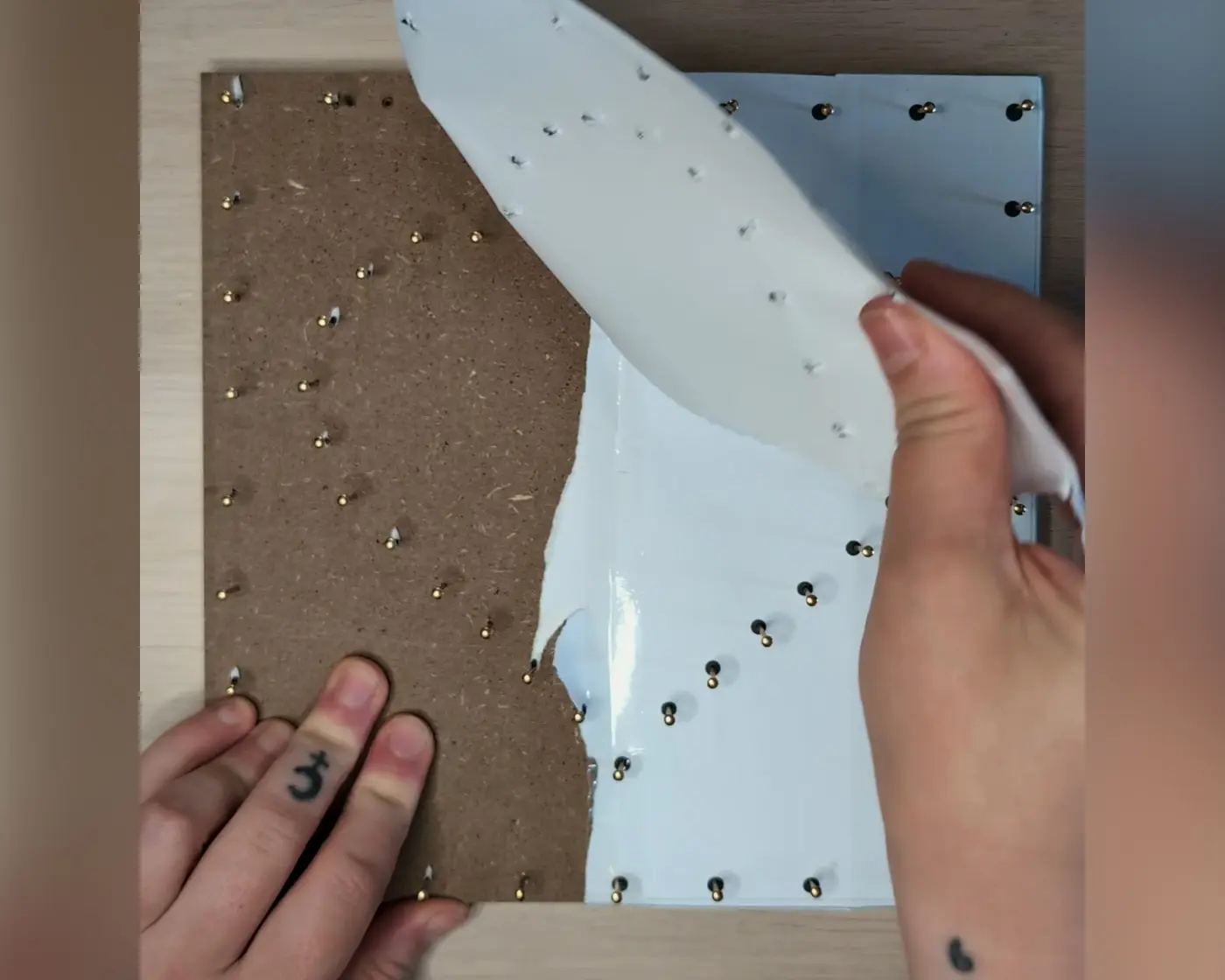 Twee handen die het papieren patroon van de houten plaat aftrekken, met de ingeslagen gouden spijkers zichtbaar