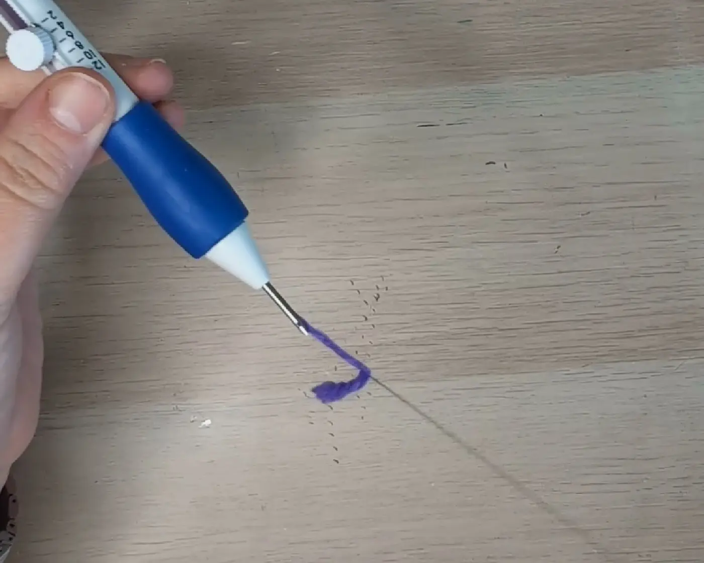 Hand houdt een blauwe punch needle waaruit paars garen naar buiten komt op een houten ondergrond