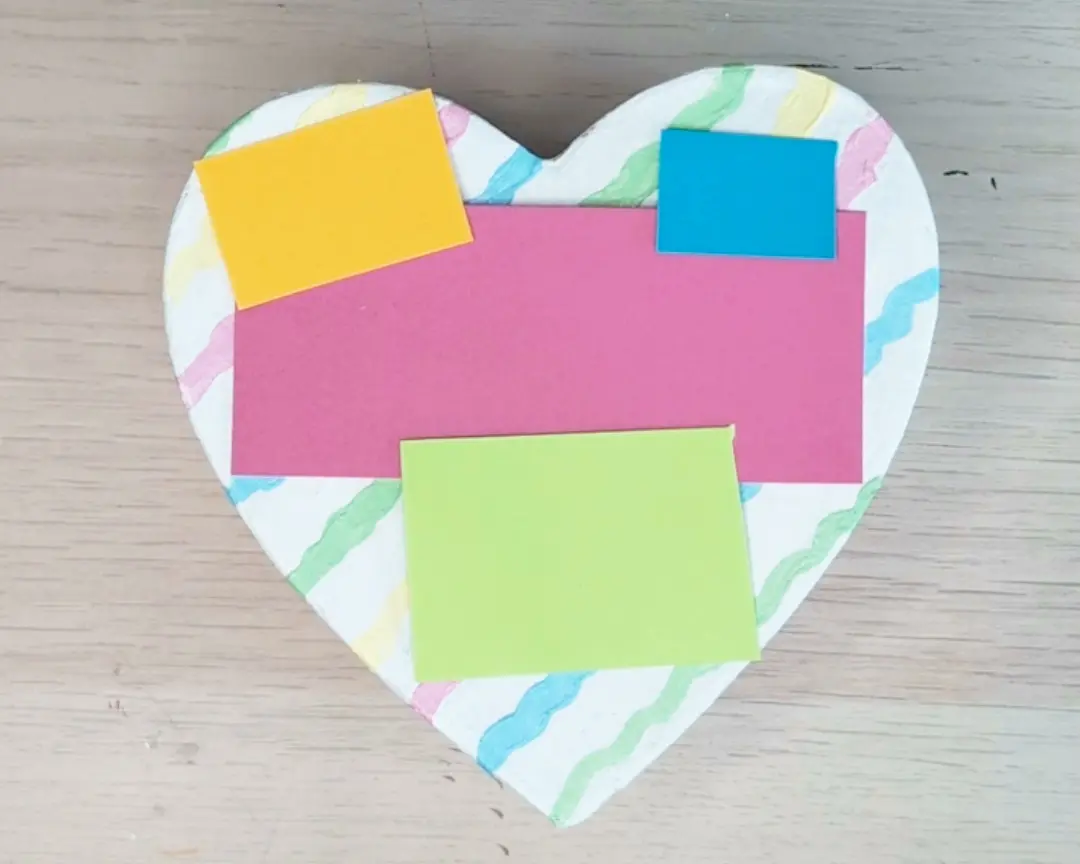 Een hartvormig deksel beschilderd met kleurrijke golvende strepen, met gele, roze, blauwe en groene gekleurde papieren rechthoeken erop gelegd als indeling.