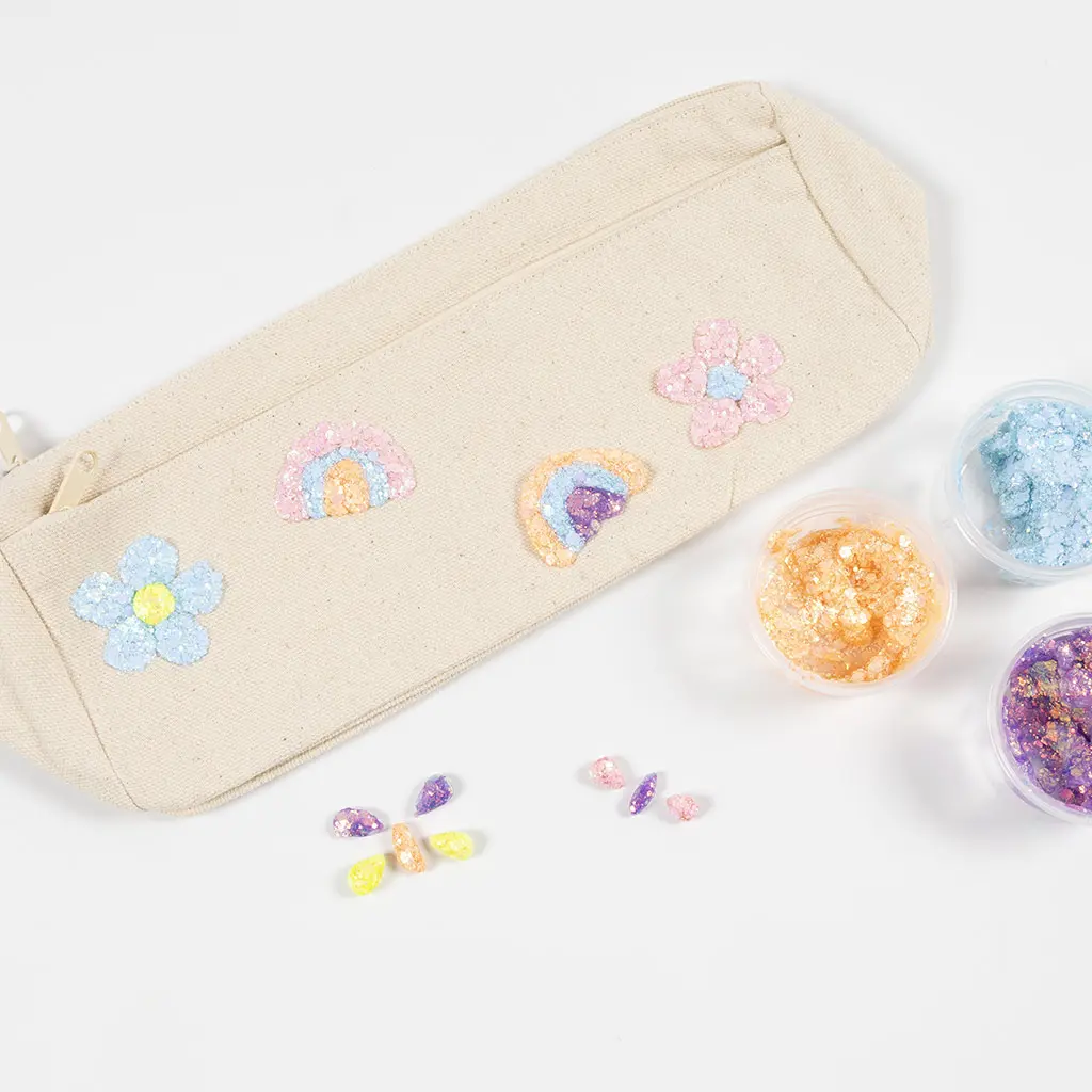 Een naturel canvas etui met glitter decoraties van een blauw bloemetje, twee regenboogjes en een roze bloemetje, naast potjes chunky glitter in goud, lichtblauw en paars, met losse glittersymbolen.
