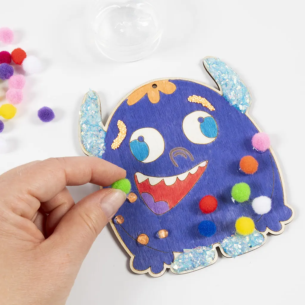 Een hand plakt een klein groen pomponnetje op het blauwe houten monster met vloeibare lijm. Er zijn al verschillende andere gekleurde pompons (zoals roze, oranje, rood en geel) op de zijkant van het monster bevestigd.