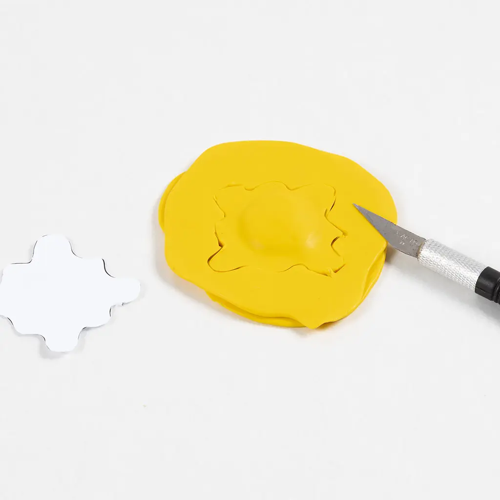 Een precisie-hobbymes snijdt een golvende vorm (zon of wolk) uit de gestapelde gele klei. Links op de witte ondergrond ligt een papieren sjabloon van dezelfde vorm.