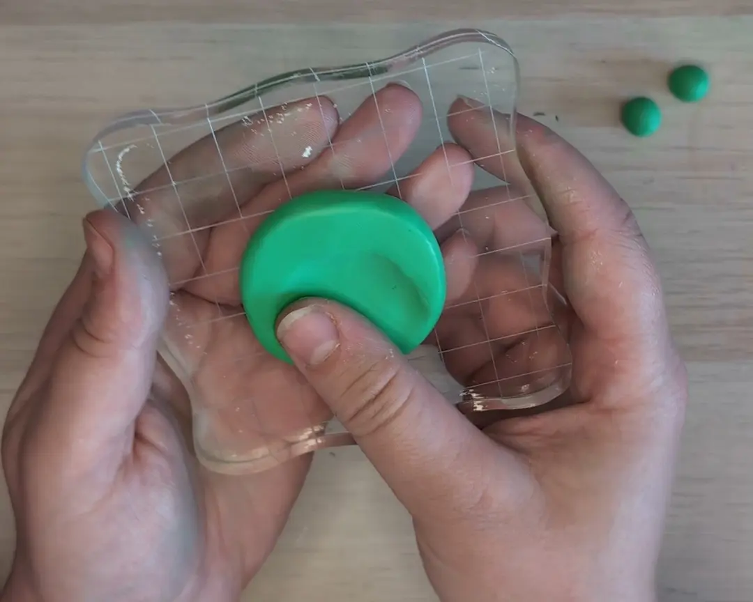 Handen houden een transparant acrylaatblok vast waarop een platgedrukte groene kleischijf ligt. Een duim drukt stevig in het midden van de schijf om een gladde holte te maken.