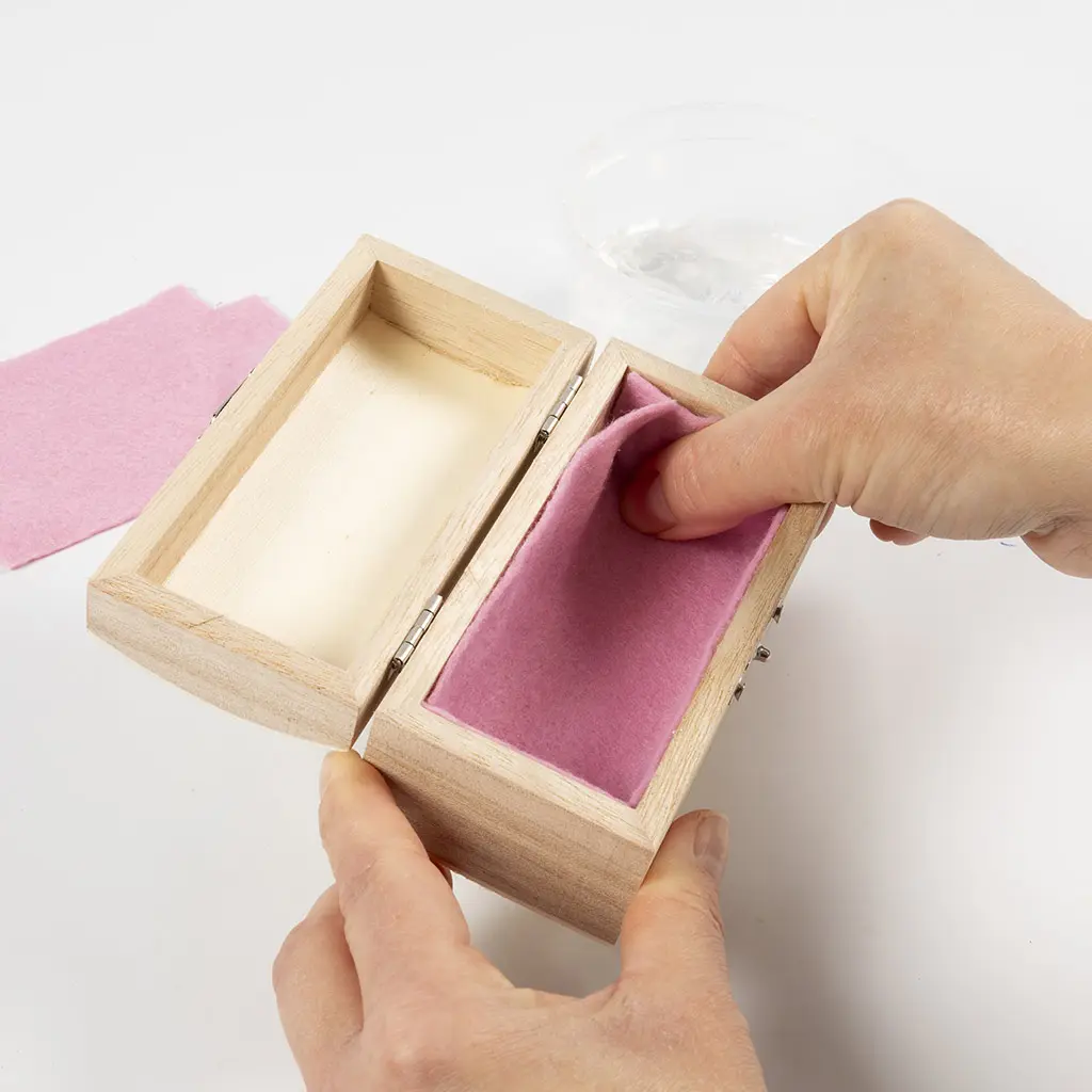 Twee handen drukken roze vilt glad tegen de binnenkant van het deksel van een open houten kistje, met losse viltstukjes en een potje lijm naast het kistje.